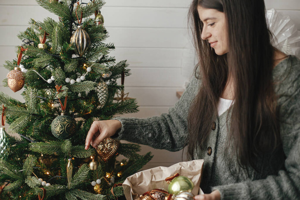 Стильная женщина в уютном свитере украшает рождественскую елку стильной безделушкой в атмосфере праздничной комнаты. Счастливого Рождества! Подготовка зимних каникул. Украшение елки с винтажным колоколом - Фото, изображение