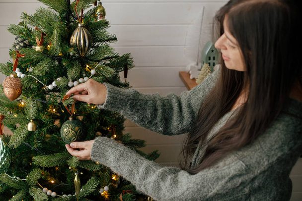 Stylish woman in cozy sweater decorating christmas tree with stylish bauble in atmospheric festive room. Merry Christmas! Winter holidays preparation. Decorating xmas tree with vintage toy - Foto, afbeelding