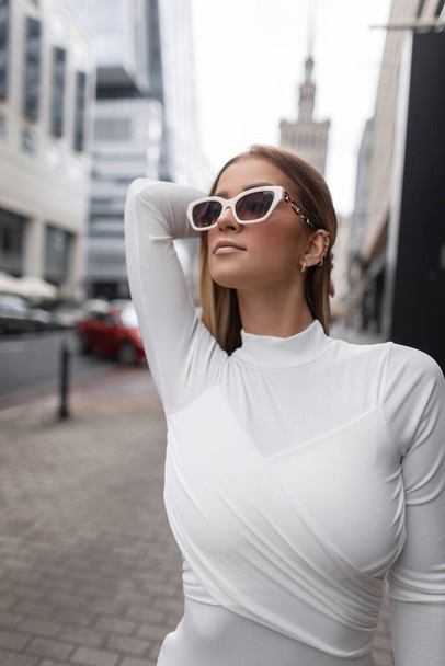 На відкритому повітрі жіночий портрет стильної красивої модної жінки з білими прохолодними сонцезахисними окулярами в білій сукні фасіони в місті
 - Фото, зображення