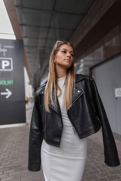 Beautiful fashionable young woman model in a stylish black leather rock jacket with a white dress walks in the city. Urban female style look outfit - Fotografie, Obrázek