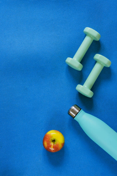 Top view of dumbbells, bottle with water and apple on blue sports mat. Flat lay, copy space. Healthy lifestyle, home training concept. - Photo, Image