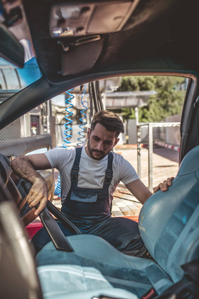 Опытный работник автосервиса в форме чистит грязное автокресло пылесосом - Фото, изображение