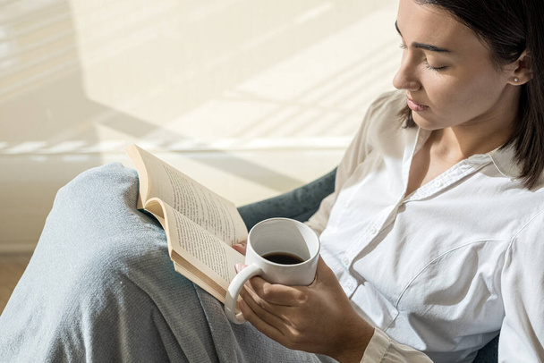 Uvolněná dívka si čte knihu s kávou v ruce. Čtení je kultura, znalosti a inteligence. Dělá to člověka více intelektuálním.. - Fotografie, Obrázek