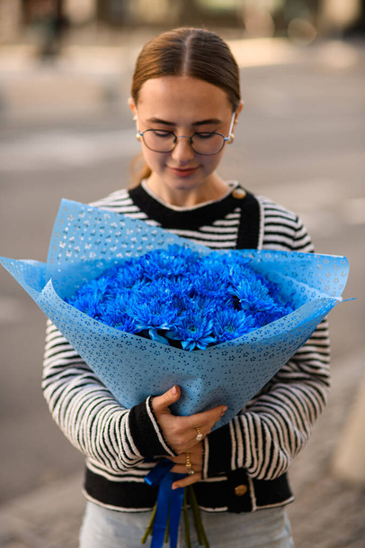 front view of handsome woman holding in her hands large lush bouquet of blue chrysanthemums in wrapping paper and looking at it - Zdjęcie, obraz