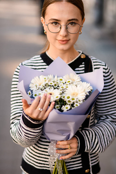 handsome woman gently holds beautiful bouquet of white flowers gerbera chrysanthemum and chamomile in wrapping paper in her hands. Close-up - Фото, изображение