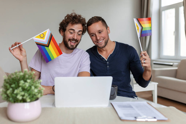 Männliche Liebhaber rufen ihre Freunde per Video im heimischen Wohnzimmer an. Ein junges schwules Paar lächelt fröhlich, während es seine Freunde per Videoanruf begrüßt. LGBTQ-Fahnen hochhalten und gemeinsam den Monat des Stolzes feiern - Foto, Bild