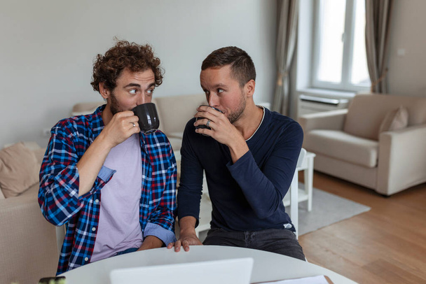 Lovely gay couple laughing together while sitting in their living room at home. Two romantic young male lovers having fun surfing the internet indoors. Young gay couple living together. - Photo, Image
