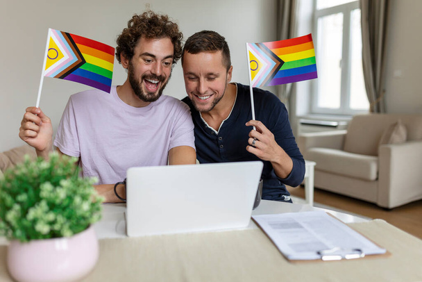 男性愛好家のビデオは自宅のリビングルームで友人を呼び出します。若いゲイのカップルは、ビデオ通話で友達に挨拶しながら陽気に笑顔を浮かべています。LGBTQの旗を掲揚し誇りを祝う月 - 写真・画像
