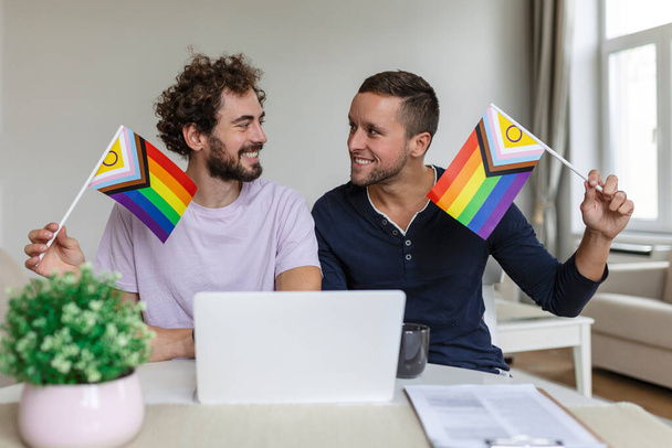 Férfi szerelmesek videó hívja a barátaikat a nappaliban otthon. A fiatal meleg pár vidáman mosolyog, miközben köszöntik a barátaikat egy videóhíváson. LMBTQ zászlók tartása és a büszkeség hónapjának megünneplése - Fotó, kép