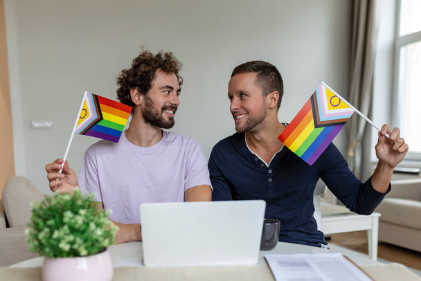 Male milenci video volá své přátele ve svém obývacím pokoji doma. Mladý homosexuální pár se vesele usmívá, zatímco zdraví své přátele na videohovoru. Držet vlajky LGBTQ a slavit měsíc hrdosti - Fotografie, Obrázek