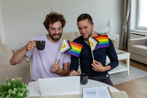 Любовники мужского пола звонят своим друзьям в гостиную дома. Молодая гей-пара весело улыбается, приветствуя своих друзей на видеозвонке. Держать флаги ЛГБТК и отмечать месяц гордости вместе - Фото, изображение