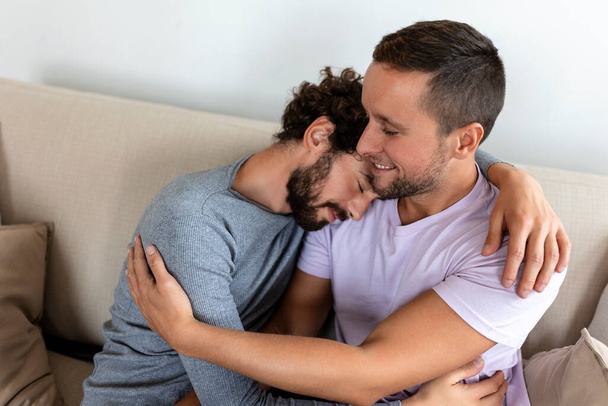 Schönes homosexuelles Paar küsst sich, während es in seinem Wohnzimmer zu Hause sitzt. Zwei romantische junge männliche Liebhaber amüsieren sich drinnen. Junge homosexuelle Paare leben zusammen. - Foto, Bild