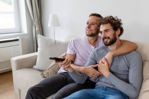 Νεαρό ομοφυλόφιλο ζευγάρι χαλαρώνει στον καναπέ στο σαλόνι, αγκαλιάζοντας και απολαμβάνοντας βλέποντας τηλεόραση - Φωτογραφία, εικόνα