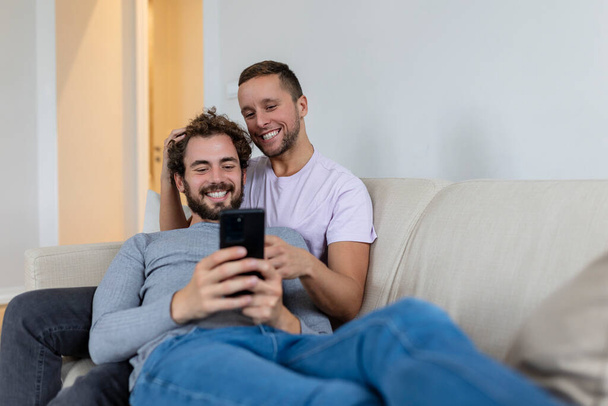 Linda pareja gay joven vídeo llamando a sus amigos en su sala de estar en casa. Dos amantes masculinos sonriendo alegremente mientras saludan a sus amigos en un teléfono inteligente. Joven pareja gay sentado juntos. - Foto, Imagen
