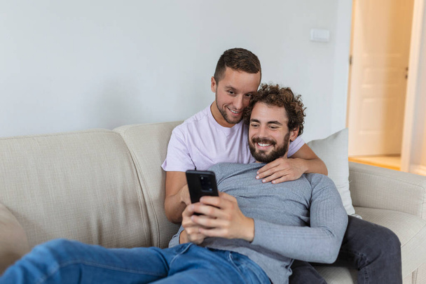 Linda pareja gay joven vídeo llamando a sus amigos en su sala de estar en casa. Dos amantes masculinos sonriendo alegremente mientras saludan a sus amigos en un teléfono inteligente. Joven pareja gay sentado juntos. - Foto, imagen