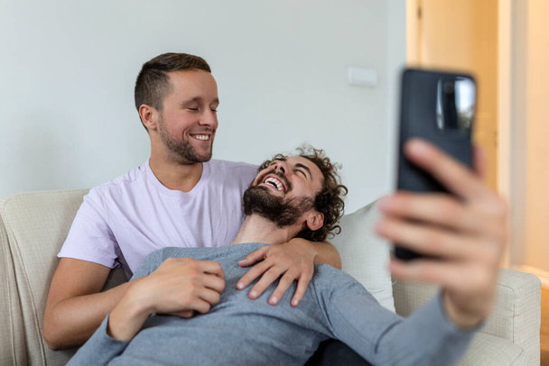 Linda pareja gay joven vídeo llamando a sus amigos en su sala de estar en casa. Dos amantes masculinos sonriendo alegremente mientras saludan a sus amigos en un teléfono inteligente. Joven pareja gay sentado juntos. - Foto, imagen