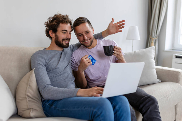 Cheerful young gay couple smiling cheerfully while shopping online at home. Two young male lovers using a credit card and a laptop to make a purchase online. Young gay couple sitting together indoors. - Fotografie, Obrázek