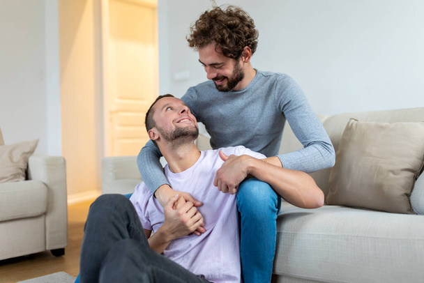 Lovely gay couple kissing while sitting in their living room at home. Two romantic young male lovers having fun indoors. Young gay couple living together. - Photo, Image