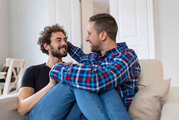 Χαρούμενο νεαρό γκέι ζευγάρι που κάθεται μαζί. Δύο στοργικοί άντρες εραστές χαμογελούν χαρούμενα αγκαλιάζοντας ο ένας τον άλλον. Νεαρό γκέι κουπέ είναι ρομαντικό. - Φωτογραφία, εικόνα