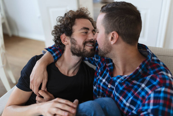 Веселая молодая гей-пара, сидящая вместе. Два любящих мужчины весело улыбаются, обнимая друг друга. Молодое гей-купе романтично. - Фото, изображение