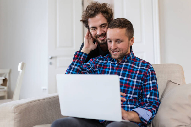 Lovely gay couple laughing together while sitting in their living room at home. Two romantic young male lovers having fun surfing the internet indoors. Young gay couple living together. - 写真・画像