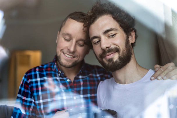 Mladý zamilovaný gay pár se dívá z okna. Dva mladí androgynní muži se spolu usmívají a dávají si kávu. - Fotografie, Obrázek