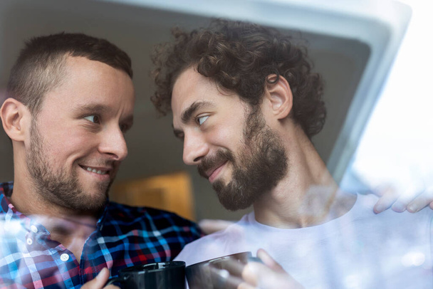 Νεαρό γκέι ζευγάρι ερωτευμένο που κοιτάει έξω από το παράθυρο. Δύο νεαροί ανδρόγυνοι άντρες χαμογελούν μαζί και πίνουν καφέ.. - Φωτογραφία, εικόνα