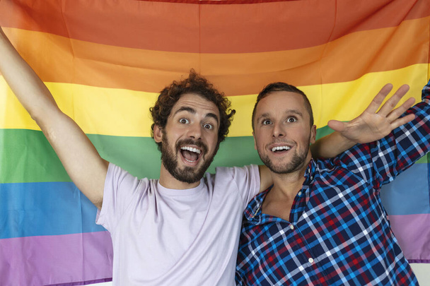 İki genç eşcinsel aşık birbirine sevgiyle bakıyor. Gurur bayrağına karşı birlikte duran iki genç erkek aşık. Sevecen genç eşcinsel çift romantik bir anı paylaşıyor.. - Fotoğraf, Görsel