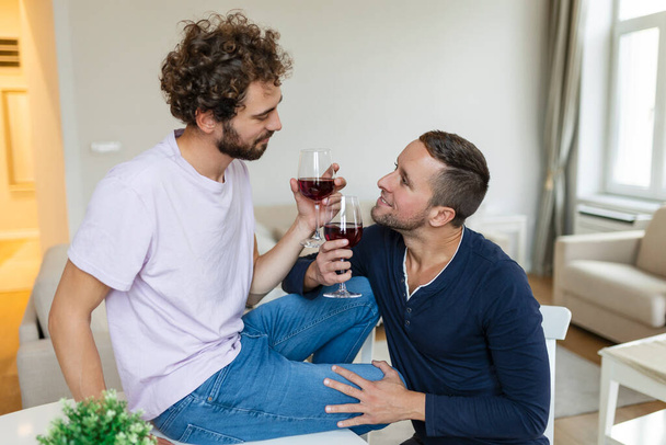 Couple LGBTQ + s'embrassant et faisant couler le vin à l'intérieur. Deux jeunes amants romantiques se regardant l'un l'autre tout en étant assis ensemble dans leur salon. Jeune couple gay étant romantique à la maison. - Photo, image