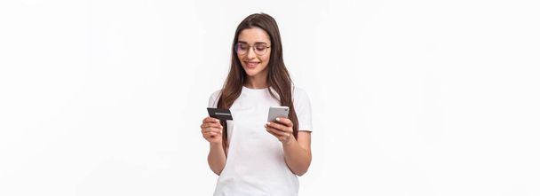Портрет привабливої молодої жінки в окулярах, що тримає кредитну картку і смартфон, введіть цифрові номери її банківського рахунку в програму покупок, замовлення онлайн, стояти на білому тлі
. - Фото, зображення