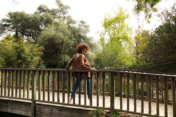 beautiful woman with curly hair leaning on the railing of a wooden bridge in a park with abundant vegetation The woman is dressed in modern and trendy clothes. Travel and holiday concept. - Zdjęcie, obraz