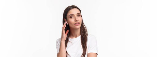 Close-up portrait of feminine attractive young woman in t-shirt, touching wireless earbud, listening music in headphone, looking camera, turn-on podcast or relaxing playlist, white background. - Photo, Image