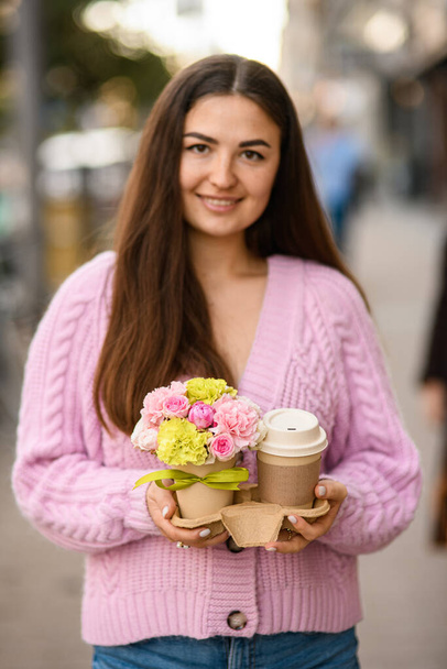 beautiful smiling woman gently holds flower arrangement of roses and carnations in small pot and paper cup of coffee in her hands. - Foto, Imagen