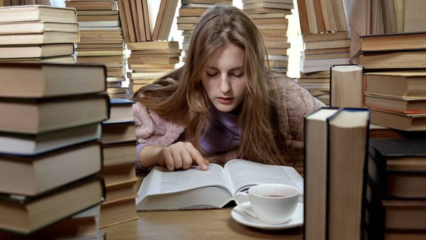 Ένα κορίτσι διαβάζει ένα βιβλίο στη βιβλιοθήκη. Στοίβες από βιβλία και τσάι στο τραπέζι. Έννοια της ανάγνωσης και της εκπαίδευσης. - Φωτογραφία, εικόνα