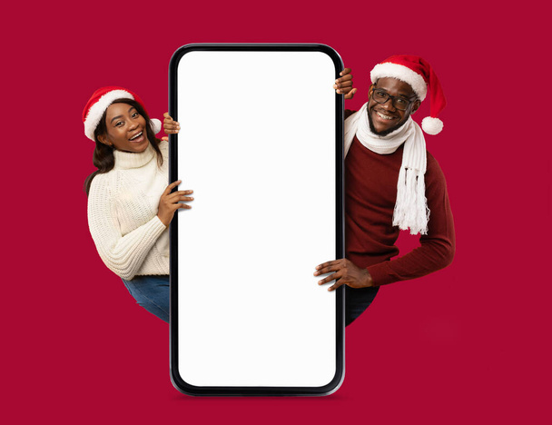 Feliz pareja de adultos negros en sombreros de Santa Claus con un gran teléfono inteligente con pantalla en blanco, recomienda para las celebraciones de Año Nuevo y Navidad, aislado en el fondo rojo, estudio. Gran oferta, anuncio y aplicación - Foto, Imagen