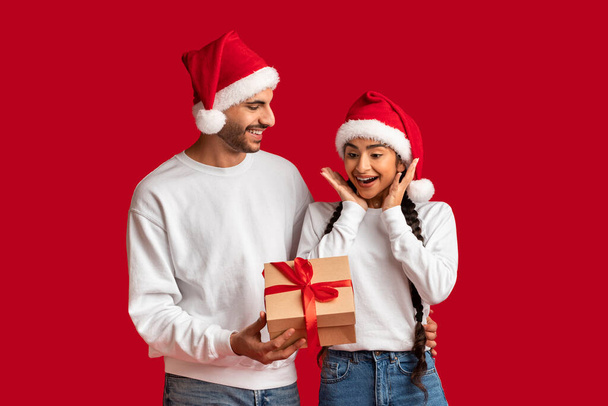 あなたへの贈り物。サンタハットのアラブ男は彼のガールフレンドにクリスマスサプライズを作ります,興奮した中東の女性にプレゼントボックスを与えます,赤の背景に隔離された立ってカップルを愛します,スペースをコピー - 写真・画像