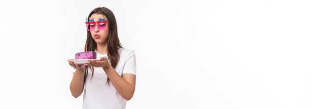 Entertainment, fun and holidays concept. Portrait of cute and funny birthday girl blowing out candle on b-day cake, wear party glasses, making wish for dream come true, white background. - Photo, Image