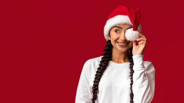 Holiday Offer. Portrait Of Cheerful Young Arab Woman Wearing Santa Hat Covering Eye With Pompom And Looking Aside At Copy Space, Happy Middle Eastern Female Posing Over Red Background, Copy Space - Photo, Image