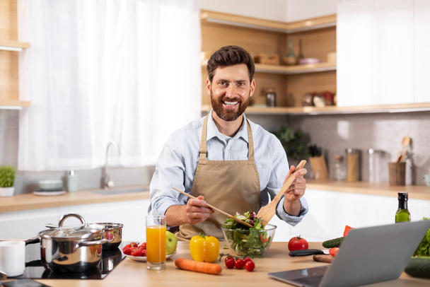 モダンなキッチンのインテリアでオーガニック野菜やノートパソコンでサラダを準備エプロンでヨーロッパのひげそりの男性シェフを笑顔。家庭での食品ブログ,家庭や食事で健康的な夕食を調理するための新しいレシピ - 写真・画像
