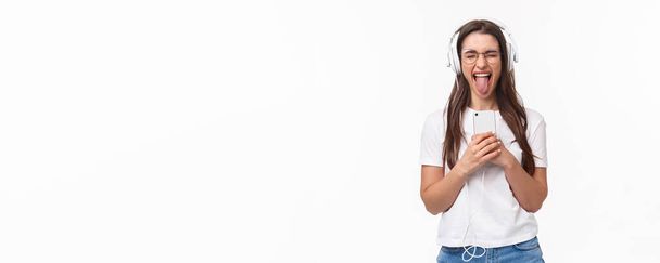 Technology, lifestyle and music concept. Portrait of carefree happy and enthusiastic woman enjoying awesome song, listening in headphones hold smartphone show tongue, white background. - Photo, Image