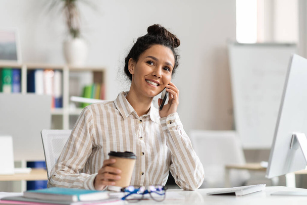 Happy female entrepreneur talking on cellphone and drinking coffee at workplace in office, smiling woman holding cup with takeaway drink and enjoying pleasant conversation - Foto, imagen