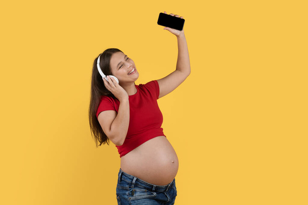 Joyeuse dame enceinte écoutant de la musique avec smartphone et écouteurs sans fil, Jeune femme enceinte heureuse tenant un téléphone mobile avec écran vide et bénéficiant d'une playlist préférée, Copier l'espace - Photo, image