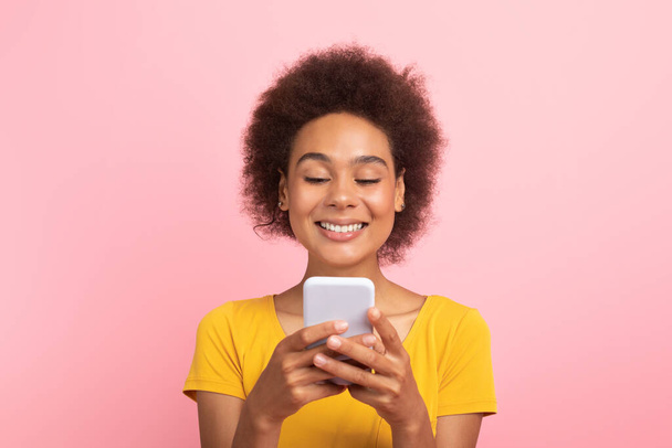 Sonriente milenaria africana americana rizada estudiante en camiseta amarilla charlando en el teléfono inteligente, aislado en el fondo rosa, estudio. Búsqueda de vídeo, surf, blog en las redes sociales, anuncio y oferta - Foto, Imagen
