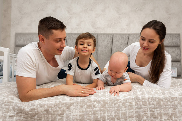 Μια μεγάλη όμορφη Ουκρανική, συναισθηματική, χαρούμενη οικογένεια του μπαμπά, μαμά και δύο μικρά παιδιά κάθεται στο κρεβάτι, φωτεινό εσωτερικό του δωματίου. Οικογενειακή έννοια  - Φωτογραφία, εικόνα