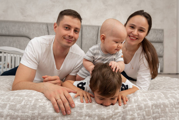Μια μεγάλη όμορφη Ουκρανική, συναισθηματική, χαρούμενη οικογένεια του μπαμπά, μαμά και δύο μικρά παιδιά κάθεται στο κρεβάτι, φωτεινό εσωτερικό του δωματίου. Οικογενειακή έννοια  - Φωτογραφία, εικόνα