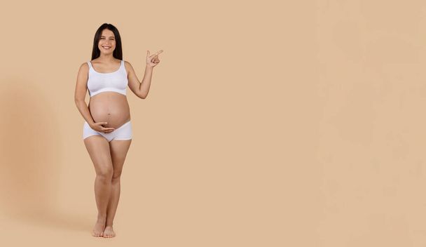 Maternity Ad. Beautiful Pregnant Lady In Underwear Pointing Aside At Copy Space, Happy Young Expectant Female Looking At Camera While Standing Over Beige Background In Studio, Panorama - Photo, Image