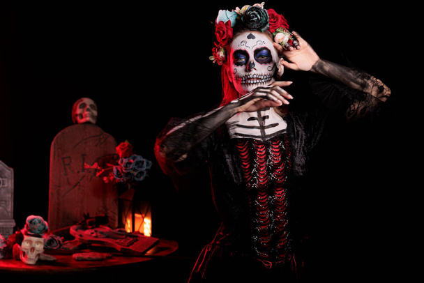 Mujer espeluznante haciéndose pasar por dama de la muerte con flores corona y cráneo maquillaje, usando traje negro de santa muerte. Celebrando dios de los muertos con body art, tradición festiva. - Foto, imagen