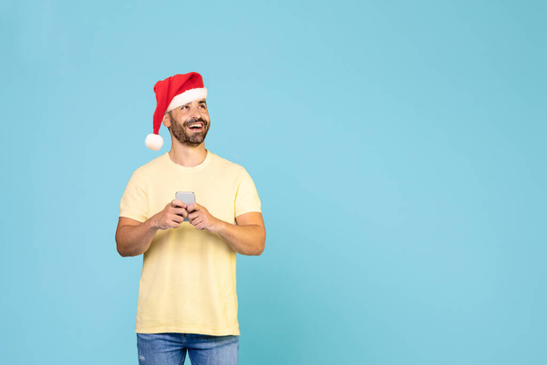 Caucasian guy in a Christmas hat with a smartphone looks at copy space, blue studio background, panorama. High quality photo - Photo, Image