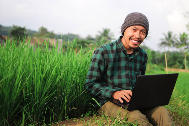 Indonezyjski młody człowiek w kapeluszu z czapką i zieloną flanelową koszulą pracujący zdalnie na środku pól ryżowych. - Zdjęcie, obraz