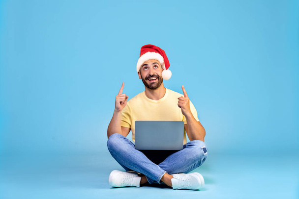 クリスマスの帽子に身を包んだ幸せな男の肖像画は、膝の上にノートパソコンを持ち、指を指している青い背景に隔離されています。高品質の写真 - 写真・画像
